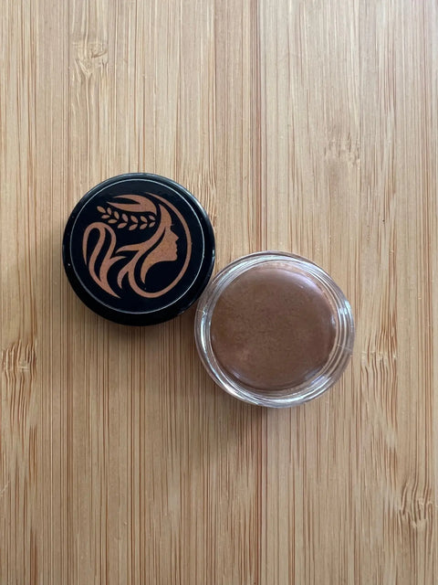 An open jar of Cheryl Kelly & Co. Triple Tint Therapy - Lip, Cheek & Eye Color with plant squalane sits on a light wooden surface. Its black lid, beside it, shows a gold profile of a woman with flowing hair and a wheat stalk.