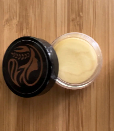 A small clear container with a black lid featuring Cheryl Kelly & Co.’s logo, filled with pale yellow Triple Tint Therapy - Lip, Cheek & Eye Color enriched with plant squalane, sits on a wooden surface.