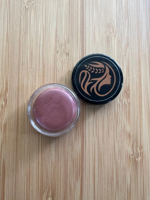 An open jar of Triple Tint Therapy - Lip, Cheek & Eye Color by Cheryl Kelly & Co. in mauve sits on a light wooden surface. The eco-friendly black lid beside it displays a stylized woman’s profile with wheat in her hair.