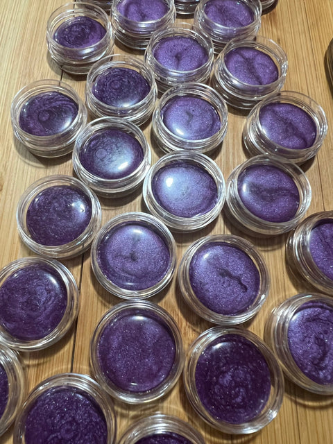 Several small, clear containers of Cheryl Kelly & Co.s Triple Tint Therapy - Lip, Cheek & Eye Color in shimmery purple are arranged on a wooden surface, highlighting the vibrant shade and eco-friendly packaging.