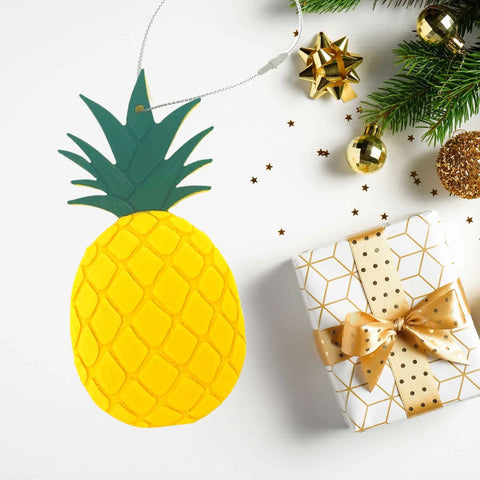 A Pineapple Christmas Ornament by Chatelet Manufacturing lies beside a gold-wrapped gift and festive pine branches. This decorative holiday ornament is made in the USA.