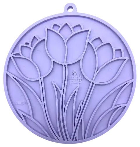 The Tulip Design EMat Enrichment Lick Mat With Suction Cups by SodaPup is a round, purple silicone mat featuring embossed tulip flowers and leaves, a hanging hole, and subtle branding. Perfect for calming your dog during snack time.