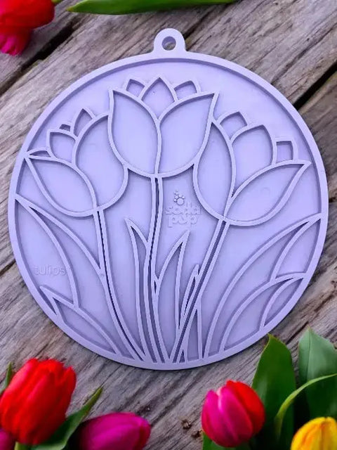 The SodaPup Tulip Design EMat Enrichment Lick Mat With Suction Cups, a round lavender mat featuring a cut-out tulip pattern, rests on a wooden surface surrounded by red, orange, and purple tulips at the corners.