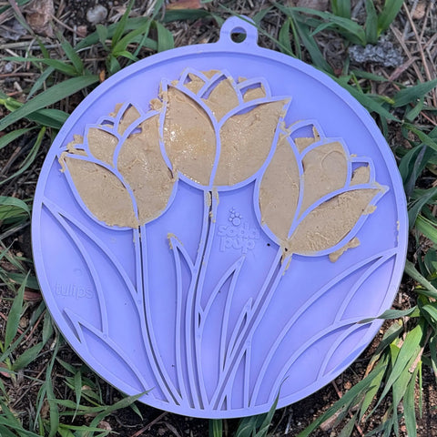 The SodaPup Tulip Design EMat Enrichment Lick Mat With Suction Cups in purple is smeared with peanut butter and placed on the grass outdoors, making it a perfect enrichment toy for pets.