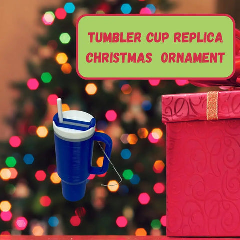 A Tumbler Cup Replica Christmas Ornament by Chatelet Manufacturing hangs before a blurred, brightly lit tree as a pink gift sits nearby. 2023s popular water bottle mug ornament—made in the USA—perfect for decorating your Christmas tree.