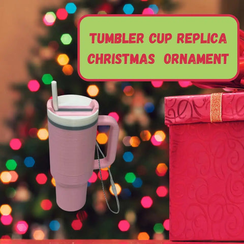 The Chatelet Manufacturing Tumbler Cup Replica Christmas Ornament—2023’s popular water bottle mug design—hangs in front of a festive tree and pink gift box, adding fun to your holiday décor. Made in the USA. Text: “Tumbler cup replica Christmas ornament.”.