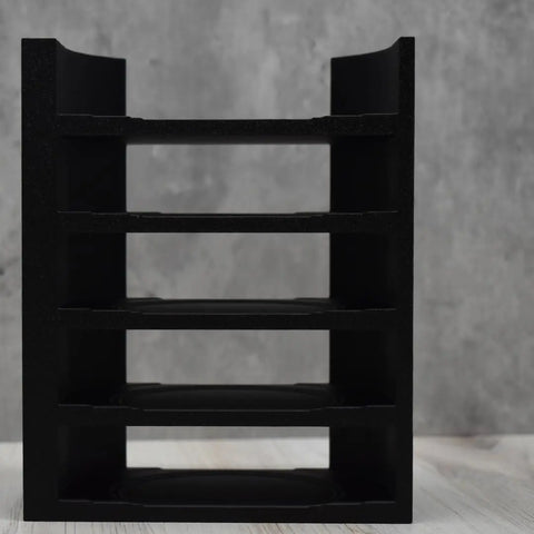 A black Chatelet Manufacturing Lid Organizer Rack—compatible with all Yeti Rambler & tumbler lids and metal straws—sits empty on a light wooden surface, front-facing against a gray, textured background. Made in the USA; fits 5 lids.