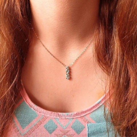 A close-up of a person with long reddish-brown hair wearing Alexa Martha Designs’ Tiny Super Sparkly Rock Candy Crystal Bar Necklace— a delicate rose gold-filled pendant with three blue stones. The person wears a pink and blue patterned top.