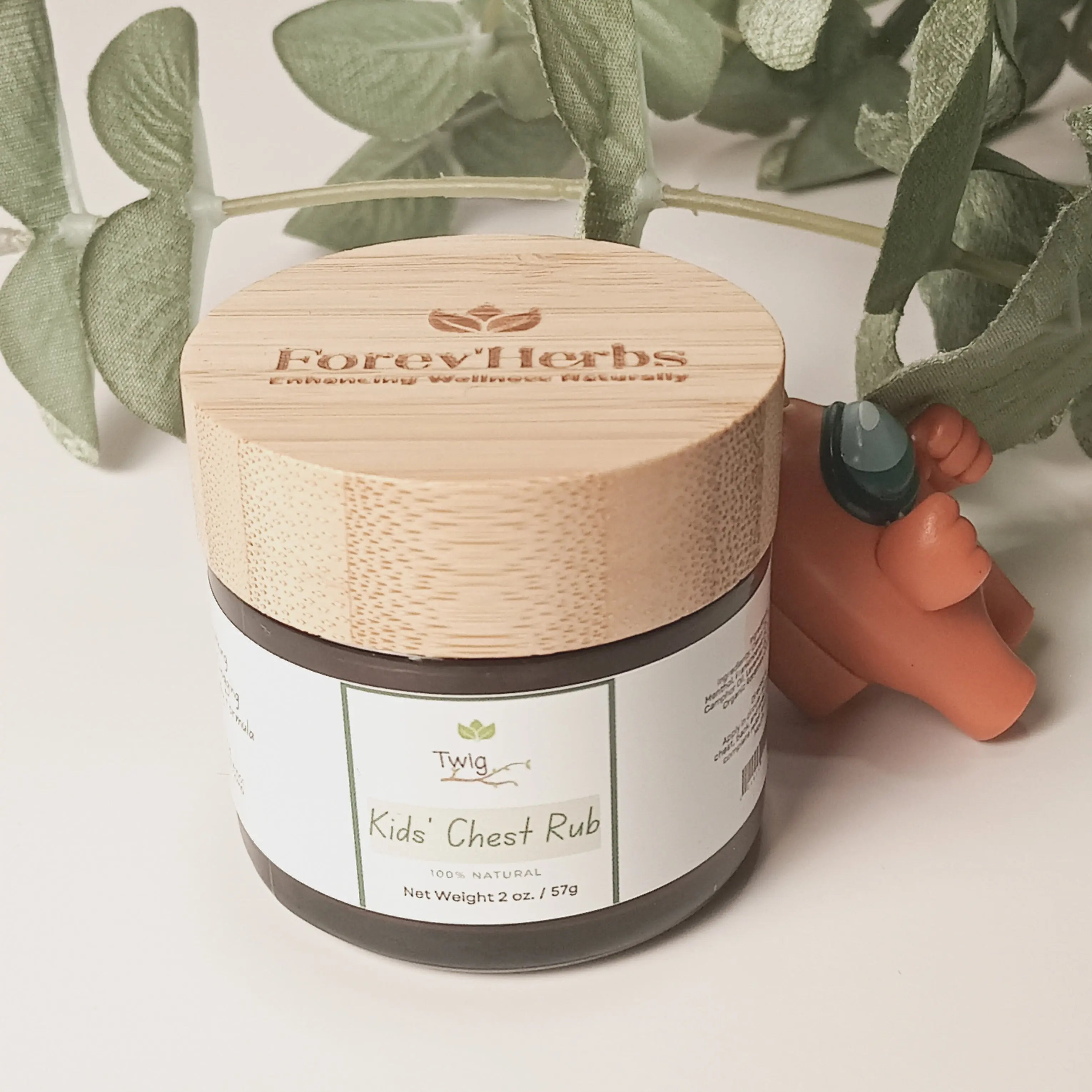 A jar of Twig - Kids' Chest Rub by Forev'Herbs with a bamboo lid sits on a white surface beside green leaves and an orange bear figurine holding a green stone.
