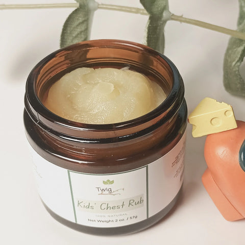 An open jar of Forev'Herbs Twig - Kids' Chest Rub with creamy balm inside sits on a white surface beside a small orange watering can, with green plant leaves in the background.