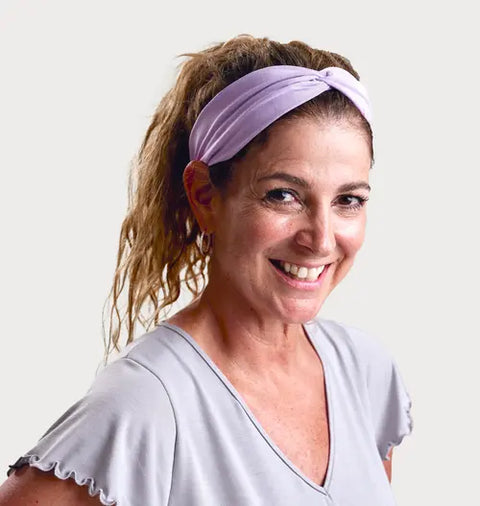 A smiling woman with curly hair wears a soft, lightweight Goodnighties Twist Headband and a light gray top, standing against a plain light background.