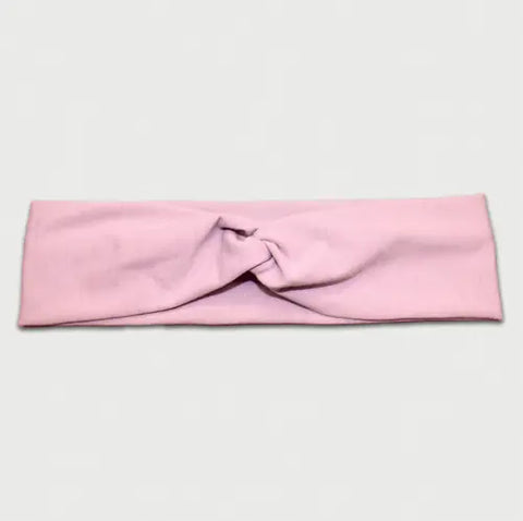 The Goodnighties Twist Headband is a soft, lightweight hair accessory in light pink with a front twist design, shown laid flat on a white background. Elegant and American-made.