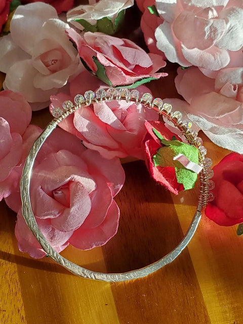 A silver, textured headband with clear crystal beads rests near a shimmering Alexa Martha Designs Two Metal Embossed Wire Wrapped Crystal Bangle on wood, surrounded by artificial pink and red roses in sunlight.