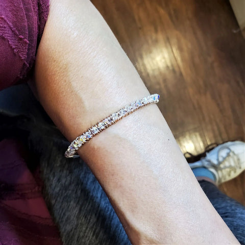 A close-up of a forearm wearing the Two Metal Embossed Wire Wrapped Crystal Bangle by Alexa Martha Designs, featuring clear stones, with wooden flooring and part of a sneaker in the background.