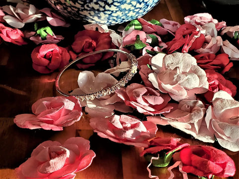 The Alexa Martha Designs Two Metal Embossed Wire Wrapped Crystal Bangle in sterling silver and copper, adorned with clear crystals, rests on wood amid artificial pink and red roses, with a blue and white patterned vase in the background.