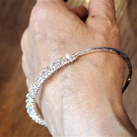 A close-up shows a hand wearing the Alexa Martha Designs Two Metal Embossed Wire Wrapped Crystal Bangle, featuring small, clear faceted beads, against a blurred brown background.