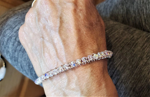 A close-up shows an elderly hand wearing the Alexa Martha Designs Two Metal Embossed Wire Wrapped Crystal Bangle, sparkling with multicolored crystals. The hand rests on a lap covered in gray fabric.