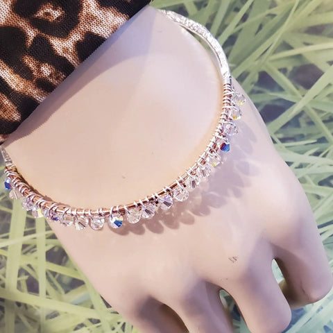 A mannequin hand displays the Two Metal Embossed Wire Wrapped Crystal Bangle by Alexa Martha Designs, featuring small clear faceted beads. The background has green, grass-like patterns and a leopard print sleeve covers part of the wrist.