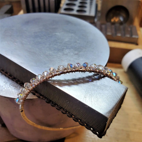The Two Metal Embossed Wire Wrapped Crystal Bangle by Alexa Martha Designs, featuring clear faceted beads, is displayed on a large metallic block in a jewelry workshop with tools in the background.