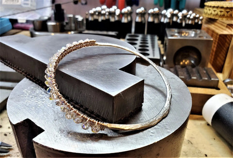 The Two Metal Embossed Wire Wrapped Crystal Bangle by Alexa Martha Designs, featuring clear beads, sits on a metal workbench in a jewelry workshop surrounded by tools and equipment.