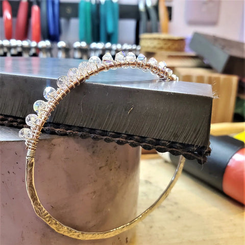 The Two Metal Embossed Wire Wrapped Crystal Bangle by Alexa Martha Designs, featuring a hammered metal band and clear beads, is displayed on a metal block in a jewelry workshop with tools visible in the background.