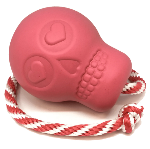 The SodaPup USA-K9 Skull Durable Rubber Chew Toy in pink features heart-shaped eyes, a stitched mouth, and a red-and-white rope, shown resting on a white surface.