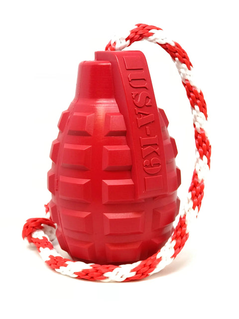The SodaPup USA-K9 Grenade Durable Rubber Chew Toy, in red with a textured surface and attached red-white braided rope, serves as a treat dispenser, tug, reward, and retrieving toy against a white background.