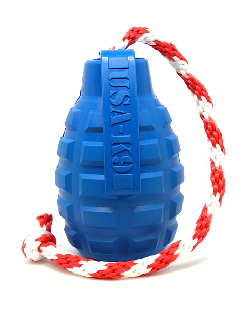The SodaPup USA-K9 Grenade Durable Rubber Chew Toy is blue, grenade-shaped with a red and white braided rope, and functions as a treat dispenser, tug, reward, and retrieving toy. Shown against a white background.