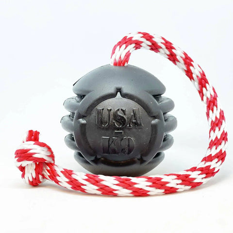The SodaPup USA-K9 Magnum Black Stars and Stripes Ultra-Durable Rubber Chew, Reward, Tug, and Retrieving Toy is shown with a red and white braided rope knotted at the end on a white background.