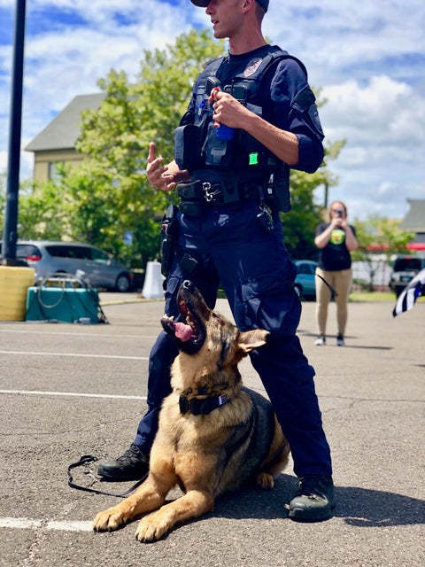 A uniformed police officer stands on pavement with a German Shepherd sitting obediently at his feet, looking up in anticipation of a reward from the SodaPup USA-K9 Magnum Grenade Durable Rubber Chew Toy and Treat Dispenser. Cars and people appear in the background.