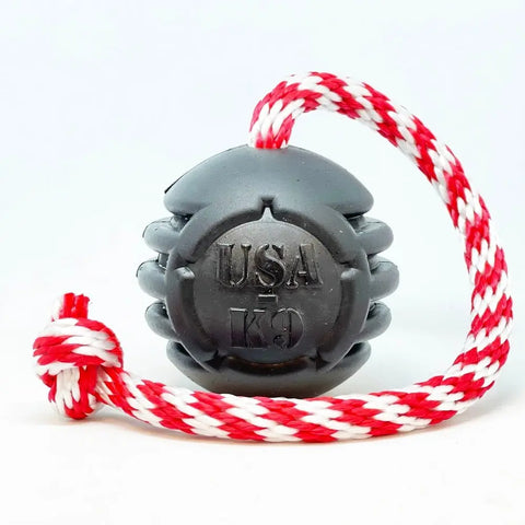 The SodaPup USA-K9 Magnum Black Stars and Stripes Ultra-Durable Reward Toy—Large Black, features embossed branding, a tough black rubber body, and a red-and-white braided rope with end knot, designed for power chewers. Set on a plain white background.