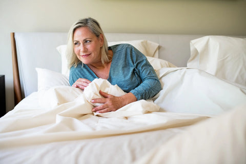 A blonde woman in a blue long-sleeve shirt lies on a bed made with the Cotton Percale Bed Sheet Set by American Blossom Linens, crafted from luxurious, USA-grown cotton, propped on one arm and smiling gently to the side.