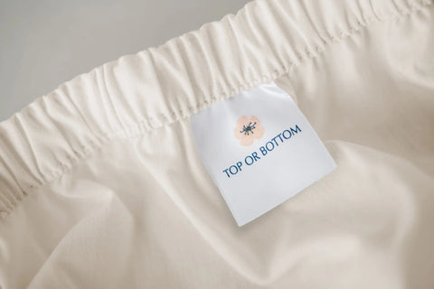 A close-up of a white fabric tag sewn into an elastic waistband of the American Blossom Linens Cotton Percale Bed Sheet Set, featuring a small flower illustration and “TOP OR BOTTOM” print on the light, off-white USA grown cotton.