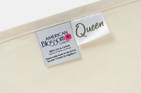 Close-up of two fabric tags on a beige textile—one says American Blossom Linens, 100% USA grown cotton, the other reads Queen—attached along the edge of these luxurious Cotton Percale Bed Sheet Sets.