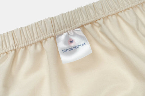 Close-up of American Blossom Linens Cotton Percale Bed Sheet Set in beige, made from USA-grown cotton, featuring an elastic waistband and a white tag reading TOP OR BOTTOM in blue with a small pink and black symbol for a luxe touch.