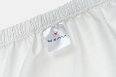 A close-up of a Cotton Percale Bed Sheet Set by American Blossom Linens, crafted from luxurious fabric, with an elastic edge and a sewn-in label reading TOP OR BOTTOM beneath a small pink flower graphic.