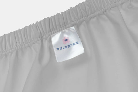 A close-up of an American Blossom Linens Cotton Percale Bed Sheet Set, made from luxurious USA-grown cotton, featuring an elastic band and a fabric label reading “TOP OR BOTTOM” beneath a small pink flower graphic.