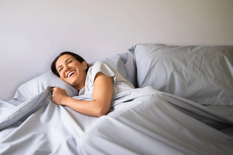 A woman relaxes in bed, smiling under the American Blossom Linens Cotton Percale Bed Sheet Set, enjoying the light gray sheets in a cozy bedroom.