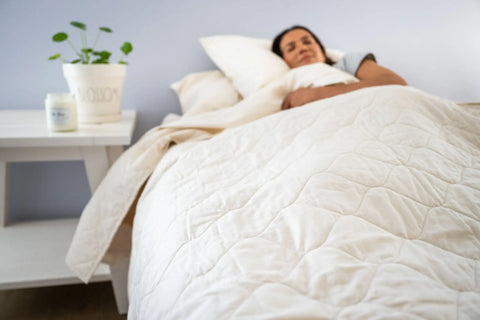 A person rests with eyes closed under the cream-colored Cotton Quilted Bedspread by American Blossom Linens. A white nightstand with a potted plant and candle completes the softly lit room, radiating eco-friendly luxury.