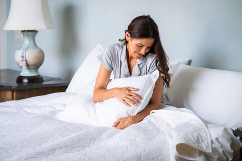 A woman in gray pajamas sits on a neatly made bed, smiling and holding a white pillow, with eco-friendly luxury Cotton Quilted Pillow Shams by American Blossom Linens and a lamp on the bedside table in the background.
