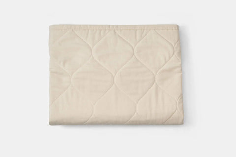 A folded beige quilted blanket with a wavy diamond pattern, crafted from USA-grown cotton, is shown on a white background. Complete the look by pairing it with American Blossom Linens Cotton Quilted Pillow Sham.