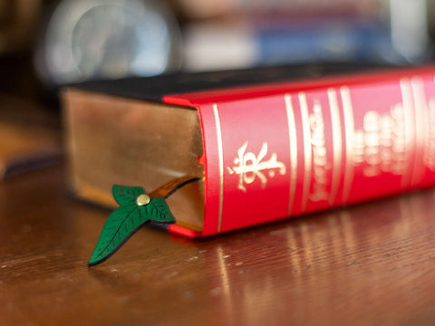 A thick red hardcover book with gold-edged pages rests on wood. A USA-Made Leather Elven Bookmark in a Lorien leaf design by KC Laser Co, partly tucked between the pages, sticks out from the book’s edge.