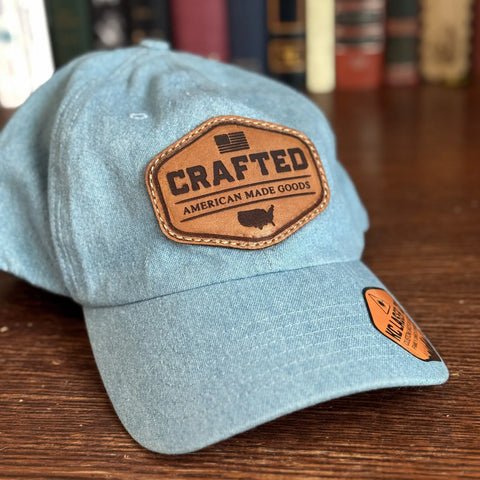 A USA-Made Solid Denim Dad Hat by KC Laser Co sits on a wooden surface, featuring a vintage leather patch with “CRAFTED AMERICAN MADE GOODS,” U.S. flag, and country outline. Blurred books in the background highlight its American craftsmanship.