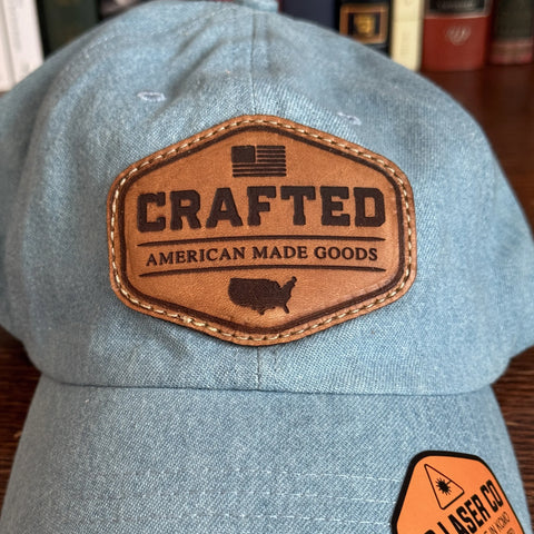 The KC Laser Co USA-Made Solid Denim Dad Hat features light blue 100% cotton, a brown leather patch with “CRAFTED AMERICAN MADE GOODS,” an American flag, and U.S. map silhouette, showcasing quality vintage American craftsmanship.