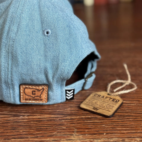 A close-up of the USA-Made Solid Denim Dad Hat by KC Laser Co shows its vintage style and leather U.S. map patch, resting on wood with a “Crafted” tag attached, highlighting handcrafted quality in the USA.