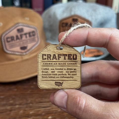 A hand holds a wooden tag reading CRAFTED AMERICAN-MADE GOODS. In the background, two KC Laser Co USA-Made Solid Denim Dad Hats with vintage leather patches, handcrafted in the USA, rest on a wooden surface.