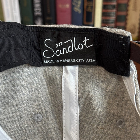 A close-up shows a black clothing label inside the gray USA-Made Solid Denim Dad Hat by KC Laser Co, reading MADE IN KANSAS CITY | USA in white, highlighting this 100% cotton vintage-style cap with leather patch. Blurred books appear in the background.
