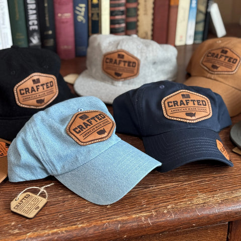 A selection of KC Laser Co USA-Made Solid Denim Dad Hats—with vintage style and front leather patch—are arranged on a wooden table. These 100% cotton caps, handcrafted in the USA, come in multiple colors. Books and more hats appear in the background.