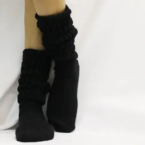 A person models Catherine Cole’s ULTIMATE Cotton Slouch Socks for women in black, standing against a white background with one leg slightly bent and crossed in front of the other.