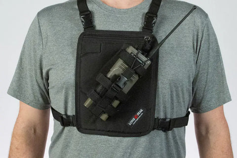 A person wearing a gray t-shirt and a Lone Peak Packs Universal Radio Chest Pack holds a handheld radio with an antenna, secured in the front pocket of the chest pack.